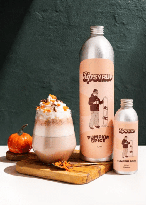 PUMPKIN SPICE SYRUP SEASON 2 a creamy hot chocolate served in a glass and topped with whipped cream and orange sprinkles next to two bottles of pumpkin spice syrup